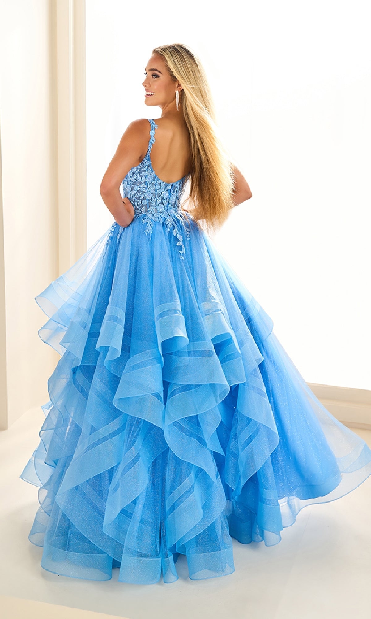 A woman stands with her back turned, wearing the Ellie Wilde EW36035 long glitter prom ball gown featuring floral bodice detailing and a sheer tulle skirt.
